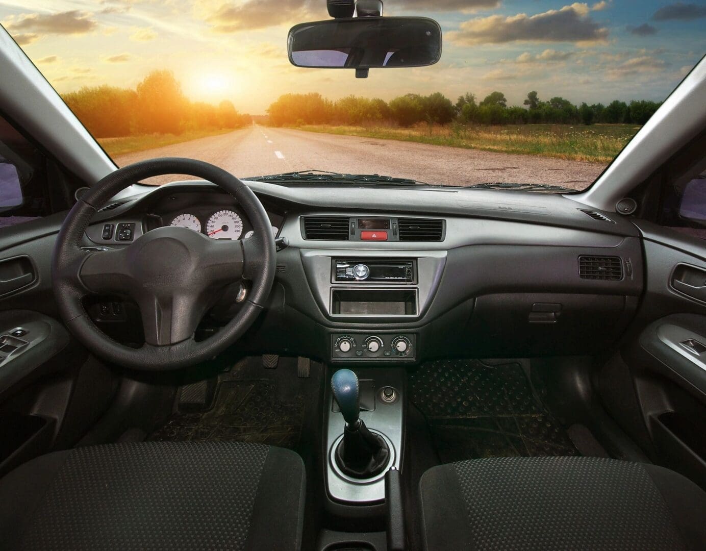 View from inside a car showing the dashboard, steering wheel, and gear shift with an empty country road and sunset visible through the windshield.