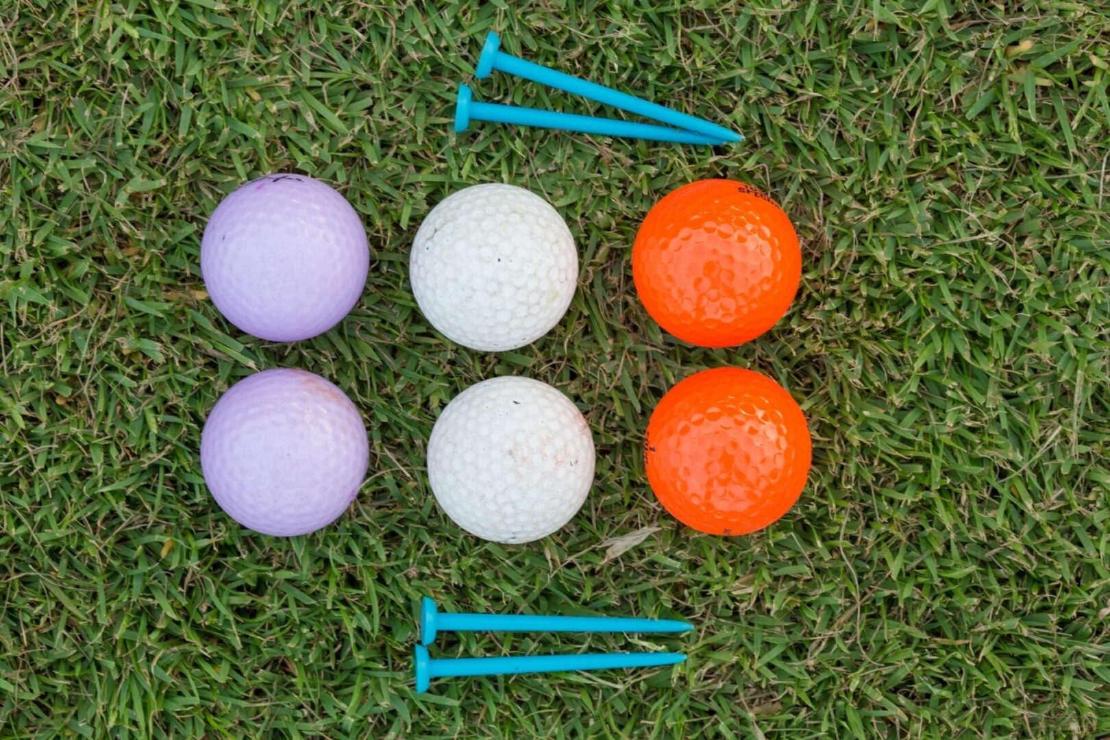 Six golf balls—two purple, two white, and two orange—are arranged in rows on grass with four blue golf tees placed above and below the balls.