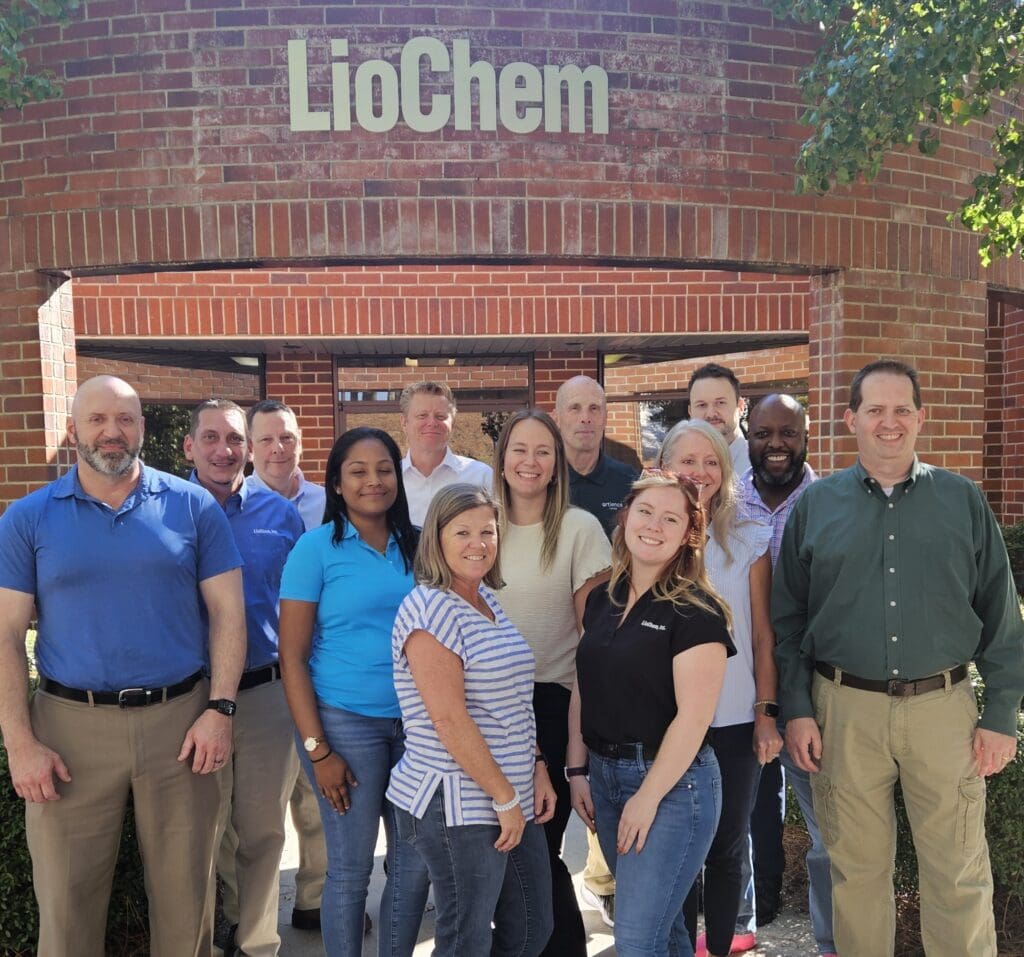 A group of twelve adults pose and smile in front of a brick building with a “LioChem” sign above the entrance.