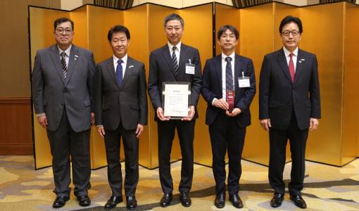 Five men in suits stand in a row indoors; the middle man holds a certificate, and the man next to him holds a red award. Gold folding screens are in the background.