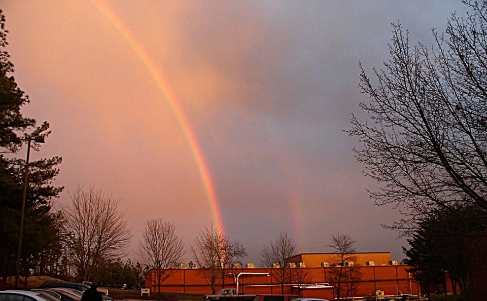 A double rainbow appears in a cloudy sky above bare trees, a brick building, and parked cars at sunset.