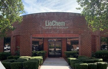 A red brick office building with a glass entrance and a sign reading “LioChem Toyo Ink Group” above the door. Shrubs line the walkway in front.