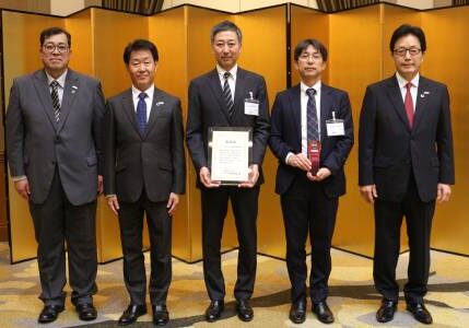 Five men in suits stand in a row indoors; the middle man holds a certificate, and the man next to him holds a red award. Gold folding screens are in the background.
